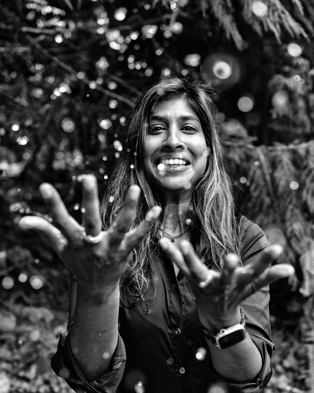 Smiling woman throwing water in open hands, standing outdoors in a wooded area, black-and-white photo