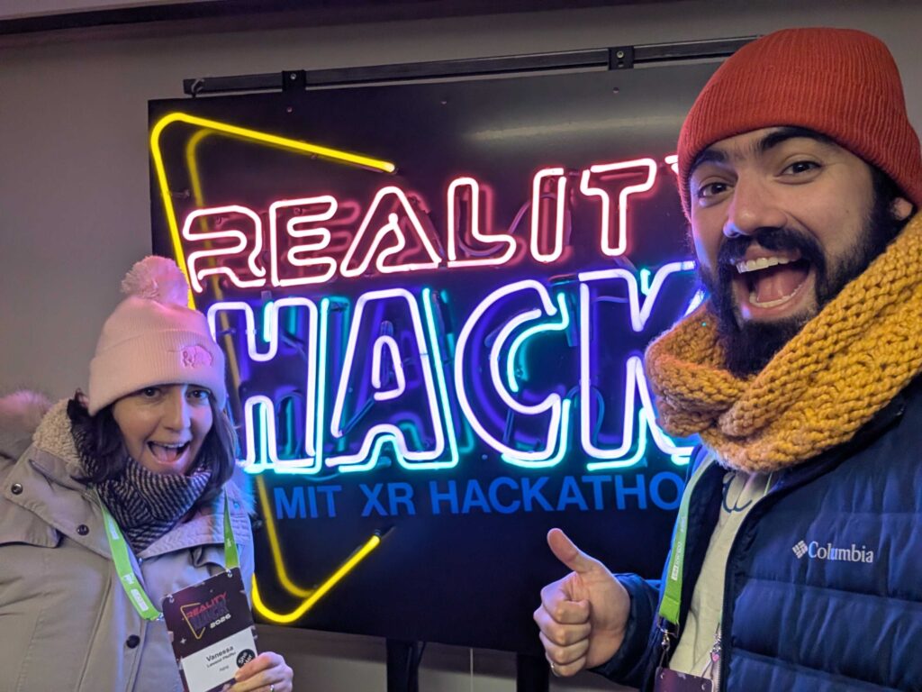 A woman and man dressed in warm close stand in front of a neon Reality Hack sign.