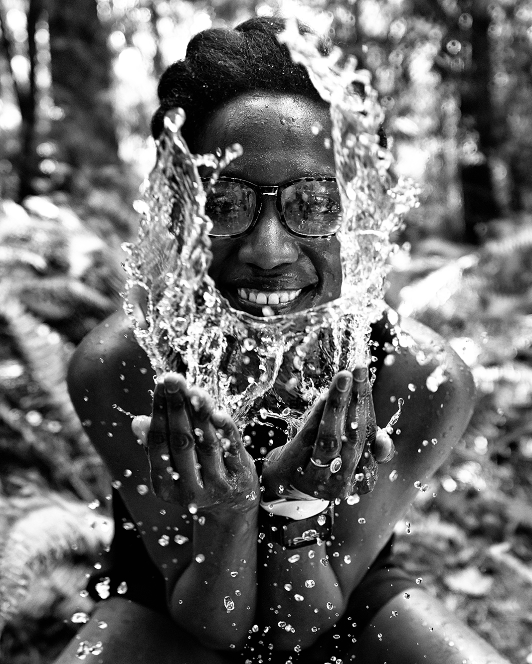 Smiling person in glasses splashing water onto their face with cupped hands, water droplets suspended midair, outdoors, black-and-white photo