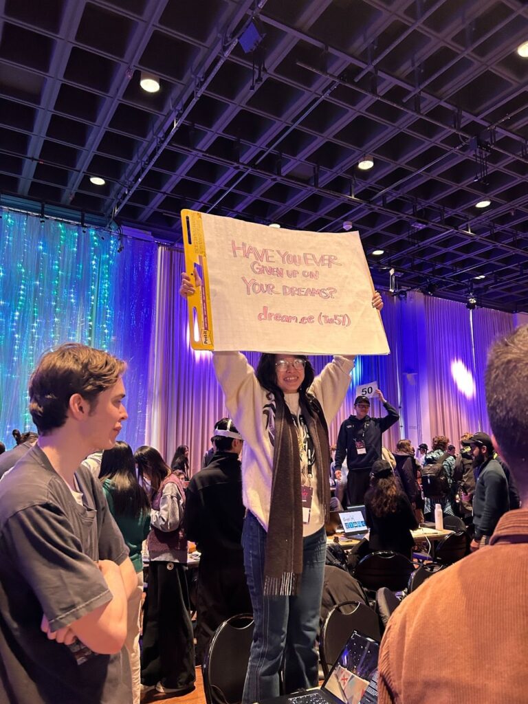 A woman stands on a chair holding a sign with the words "Have you ever given up on your dreams?"