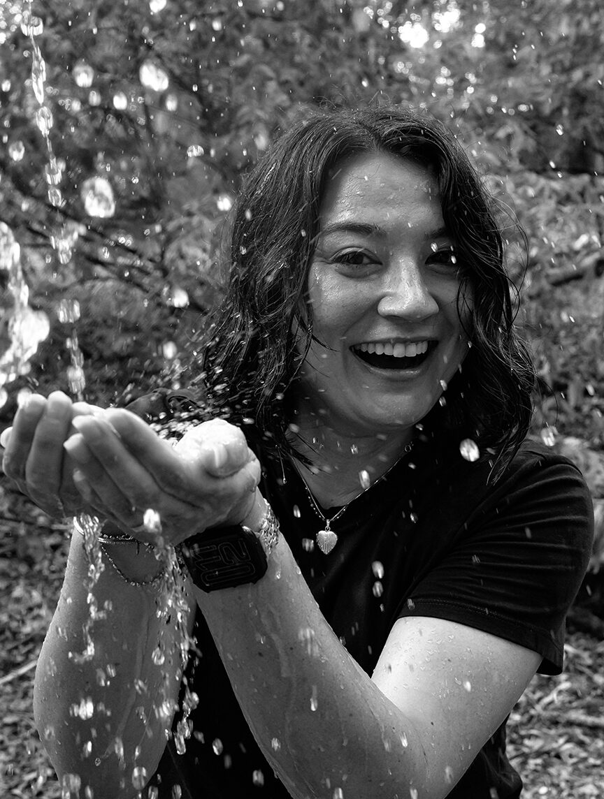 Laughing woman catching falling water in cupped hands, hair and shirt soaked, standing outdoors in a wooded area, black-and-white photo