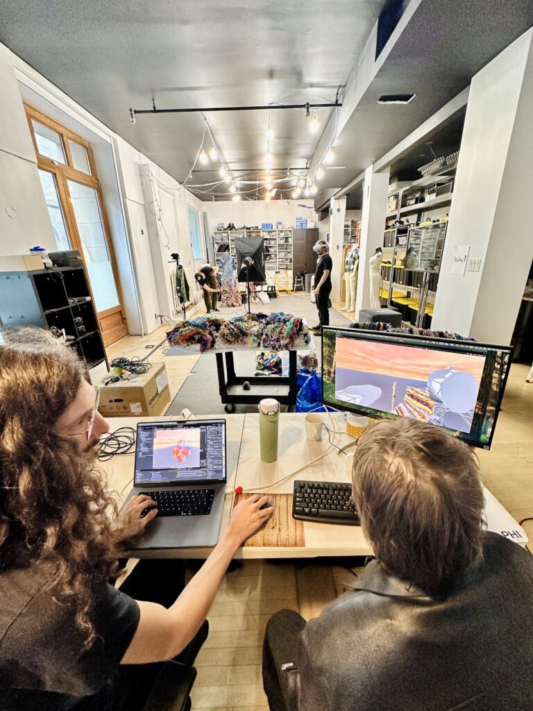 Two people sit in front of computers with digital renders of gathered cloths. In the background a person wears a headset testing the experience.