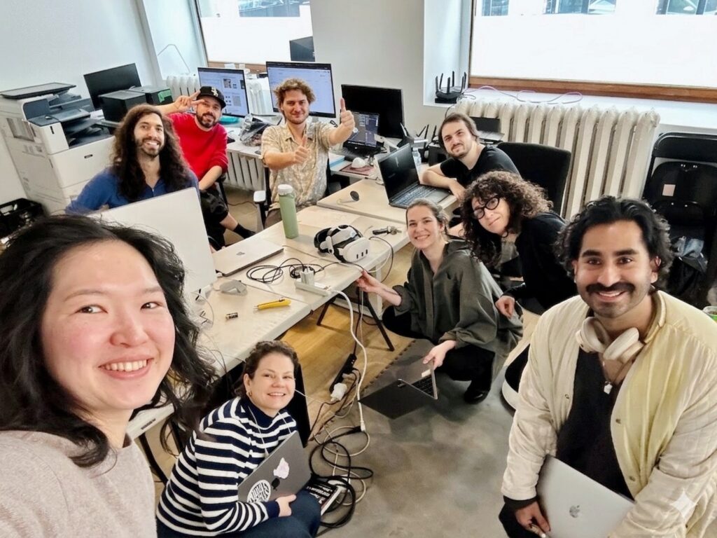 Mira takes a selfie with eight other team members in a studio filled with equipment, headsets, laptops, and monitors. 