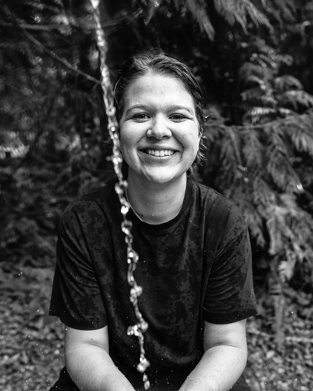 Smiling person soaked in water from an overhead stream, sitting outdoors against dark evergreen branches, black-and-white photo