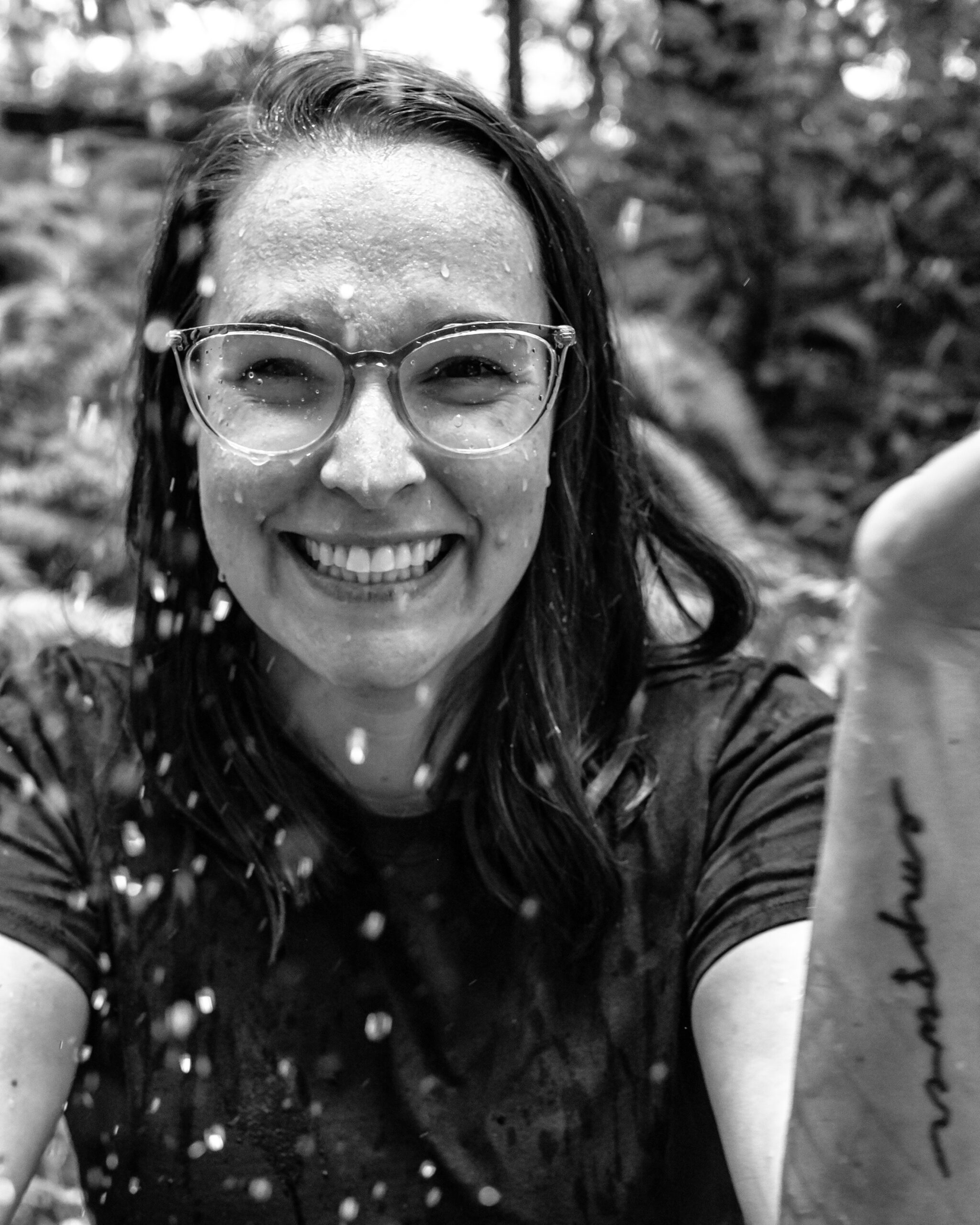 Smiling woman with wet hair and glasses, water droplets falling around her, standing outdoors in a forest, black-and-white photo
