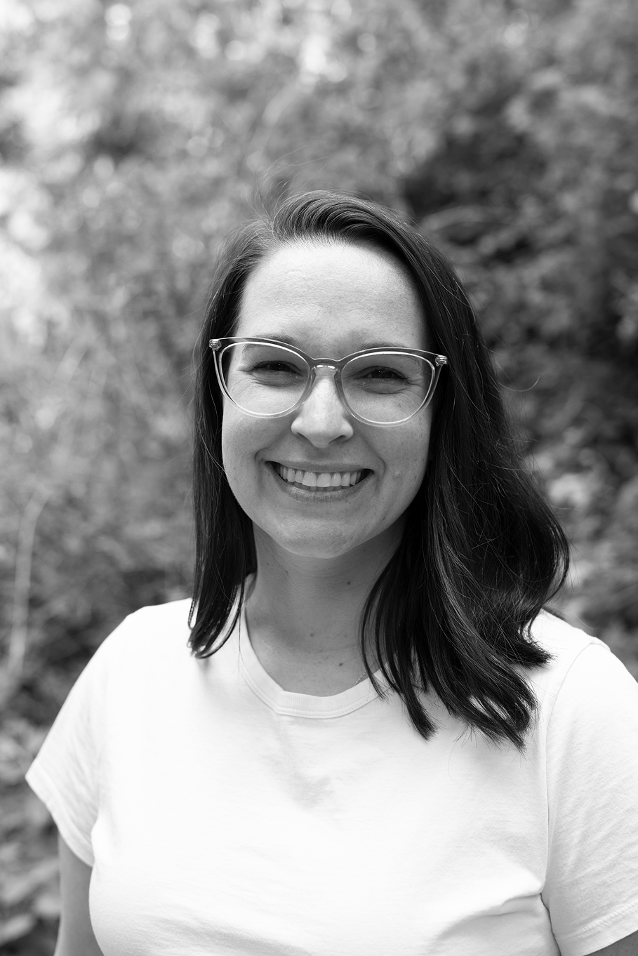 Black-and-white headshot of a smiling woman with long dark hair and glasses, wearing a t-shirt, outdoors with foliage in the background