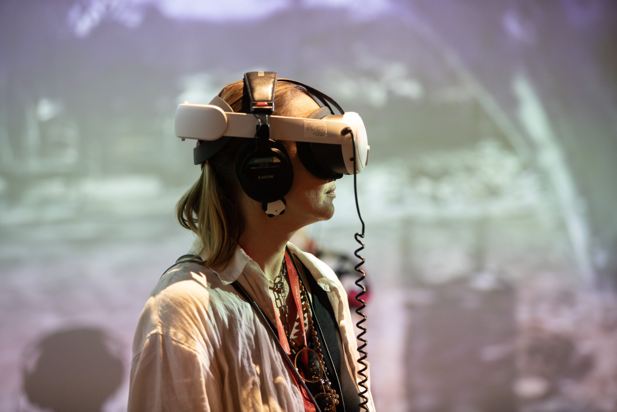 Woman in profile wearing a VR headset with wired Sony headphones and a lanyard, at an event with a blurred projected backdrop
