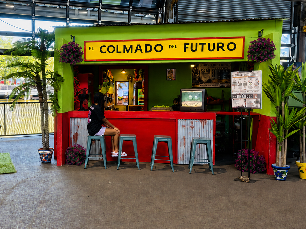 Person seated at 'El Colmado del Futuro' storefront installation with red counter, stools, potted palms, and a small TV inside