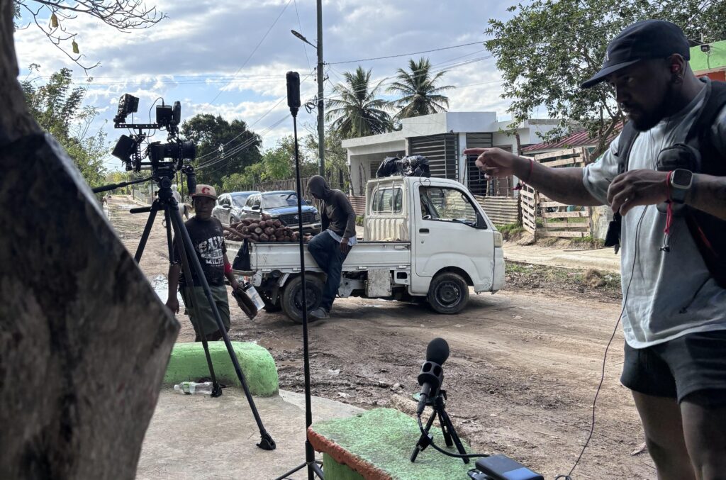 Film crew setting up camera and audio equipment on a tropical street beside a pickup truck loaded with produce