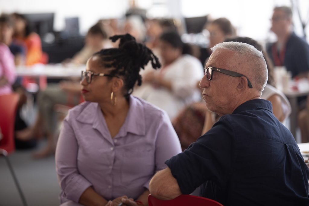 Woman in glasses and man in glasses seated side by side, looking at stage at a crowded event