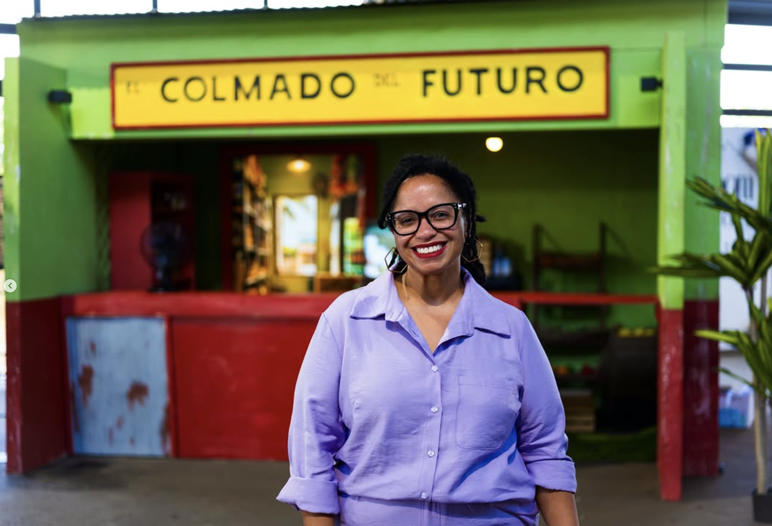 Smiling woman with glasses and red lipstick standing in front of 'El Colmado del Futuro' storefront with green and red facade