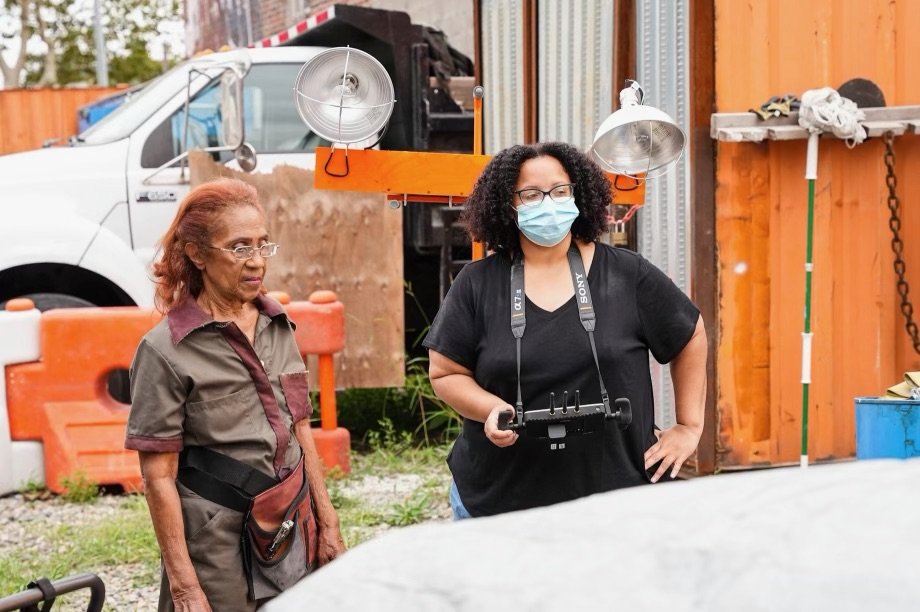 Two women on a film set, one older woman and one wearing a playback monitor and face mask