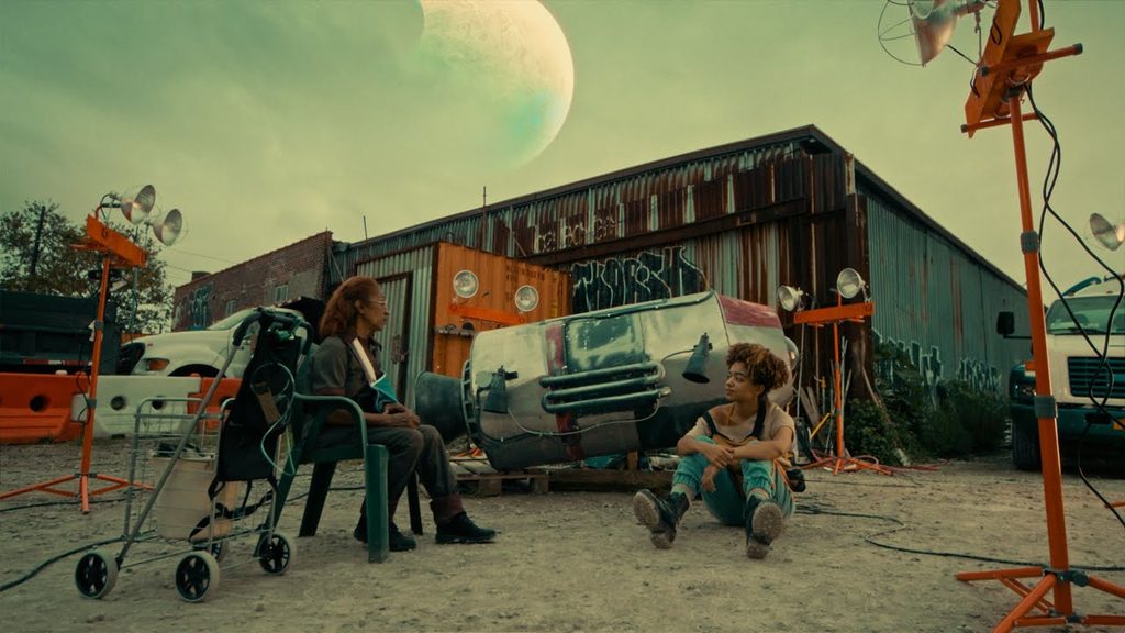 Film still: two people seated outdoors near a rusted shed and a crashed vehicle, a large planet looming in the sky above, film lights on set